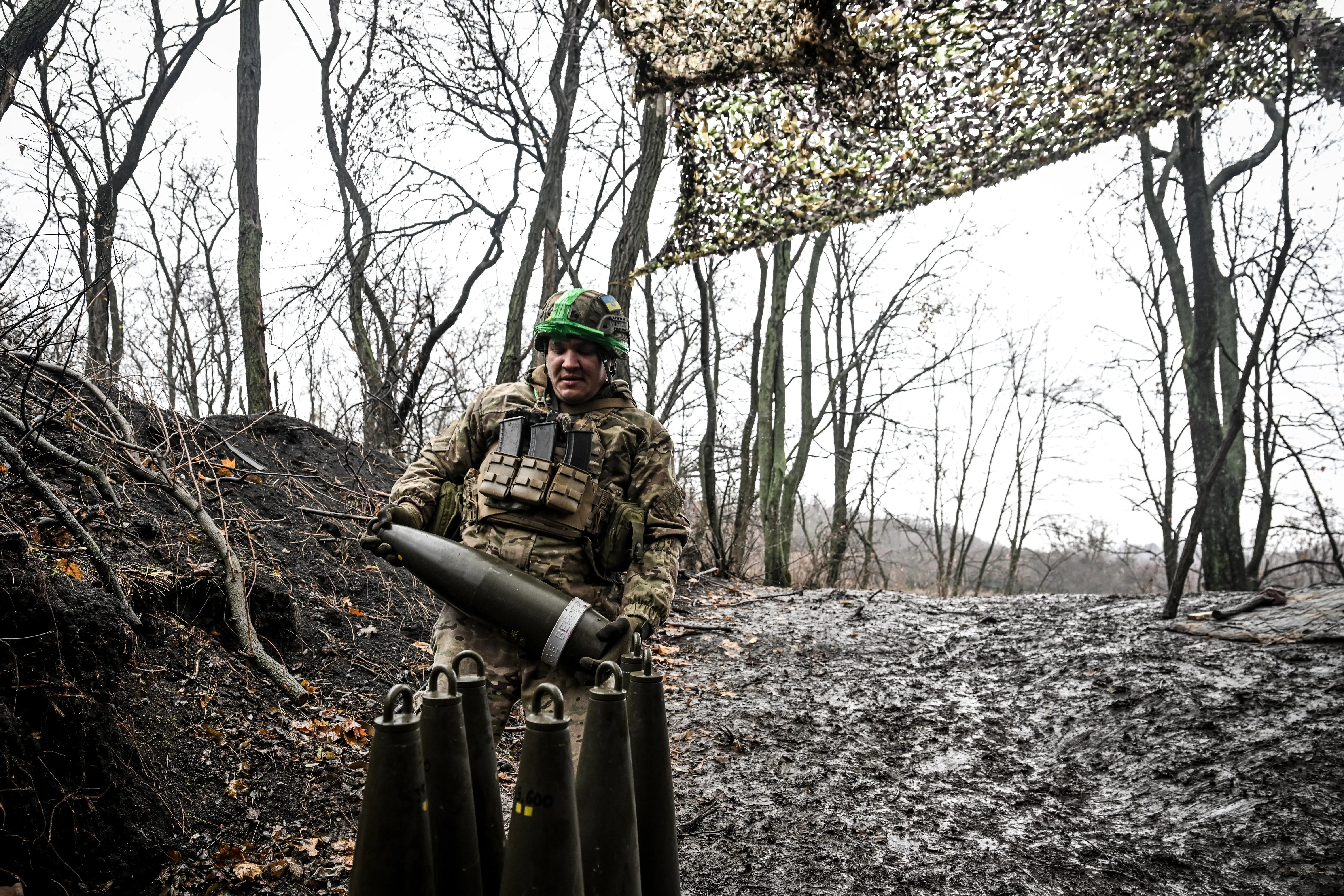 A man in camouflage stands under netting between trees under a grey sky, holding a large artillery shell