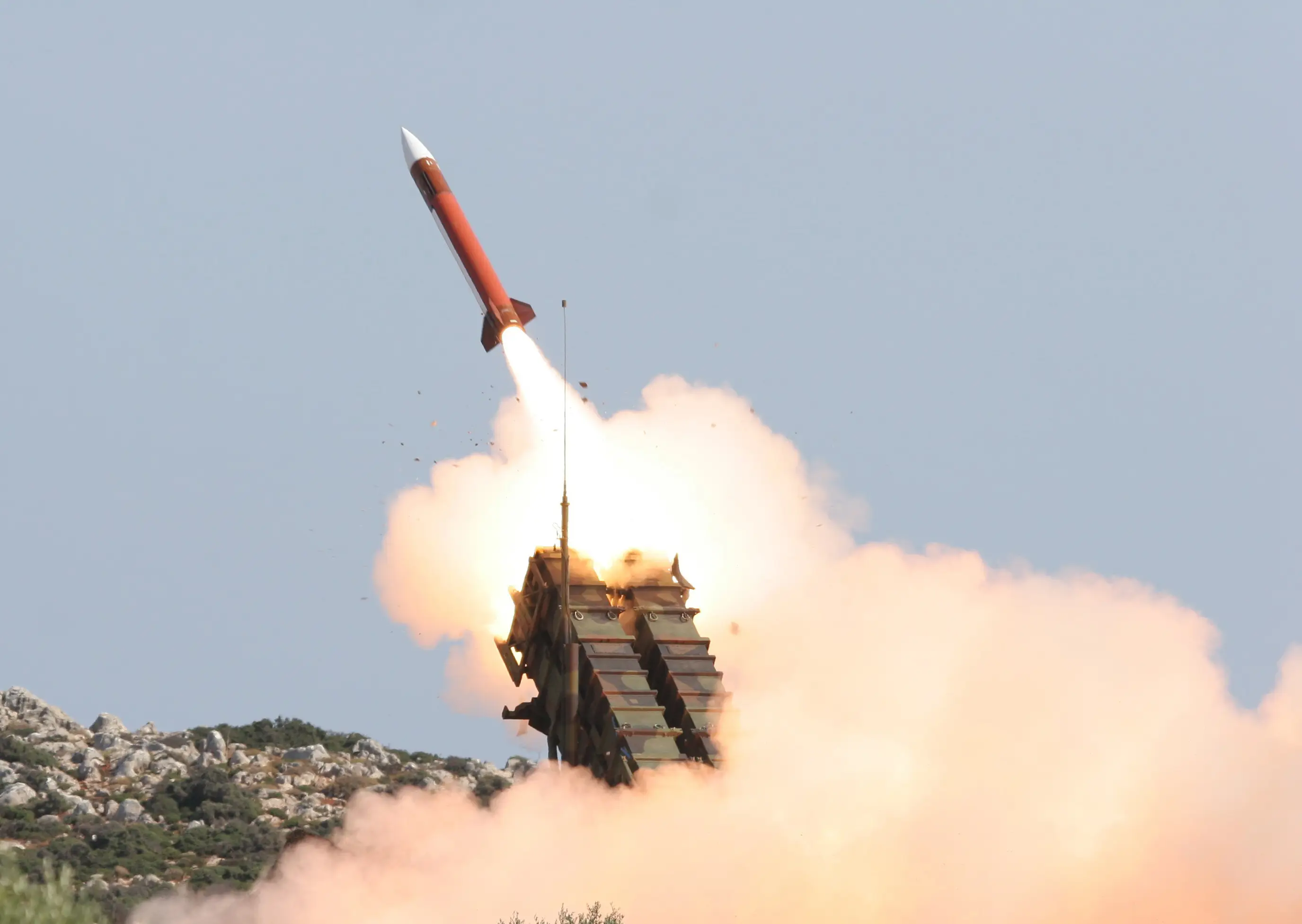 A red missile fired into the air from a camouflage-colored large weapons system, with orange smoke billowing