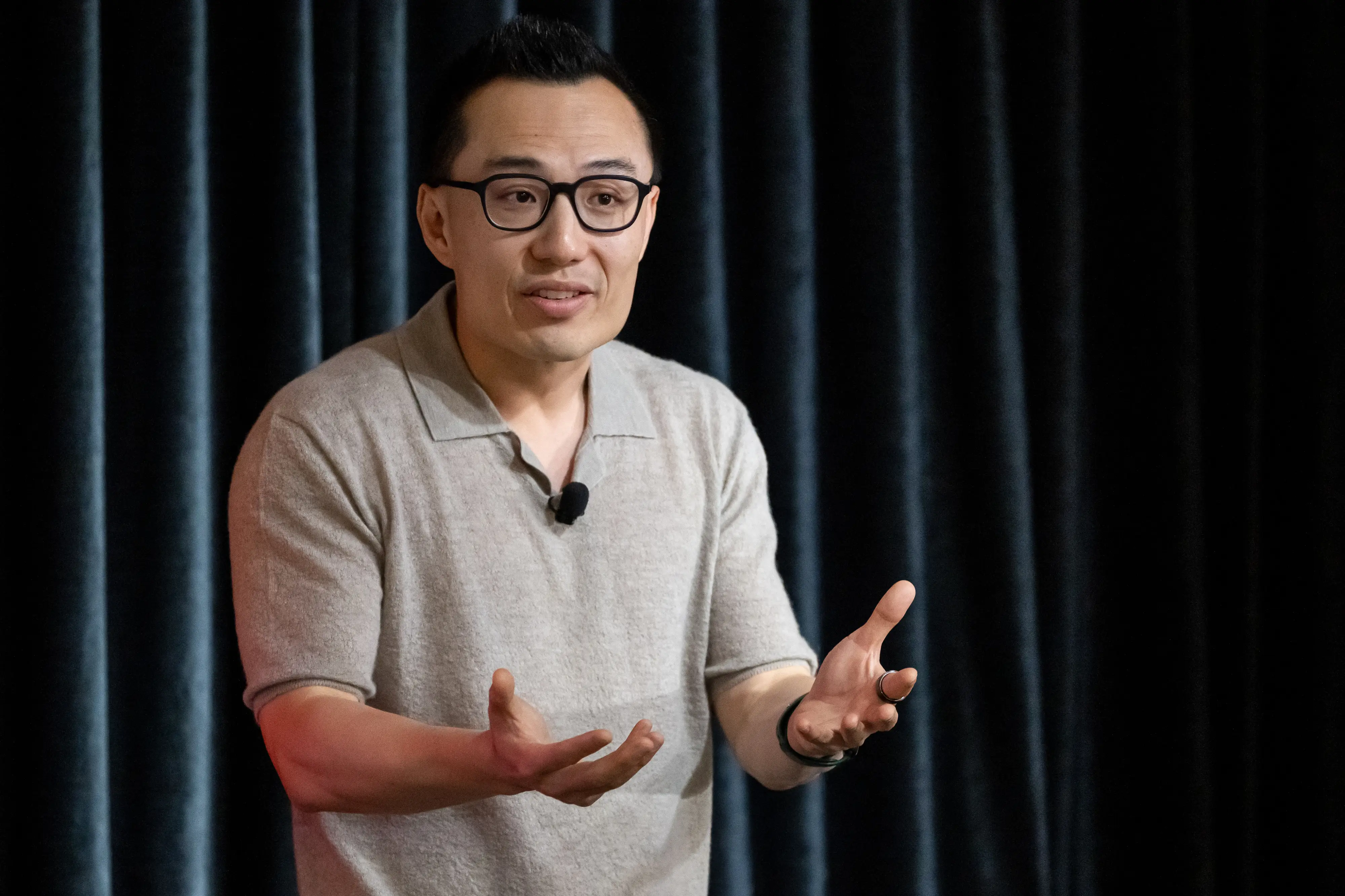 DoorDash CEO Tony Xu stands and gestures on a stage while wearing glasses with black rims and a light-grey t-shirt