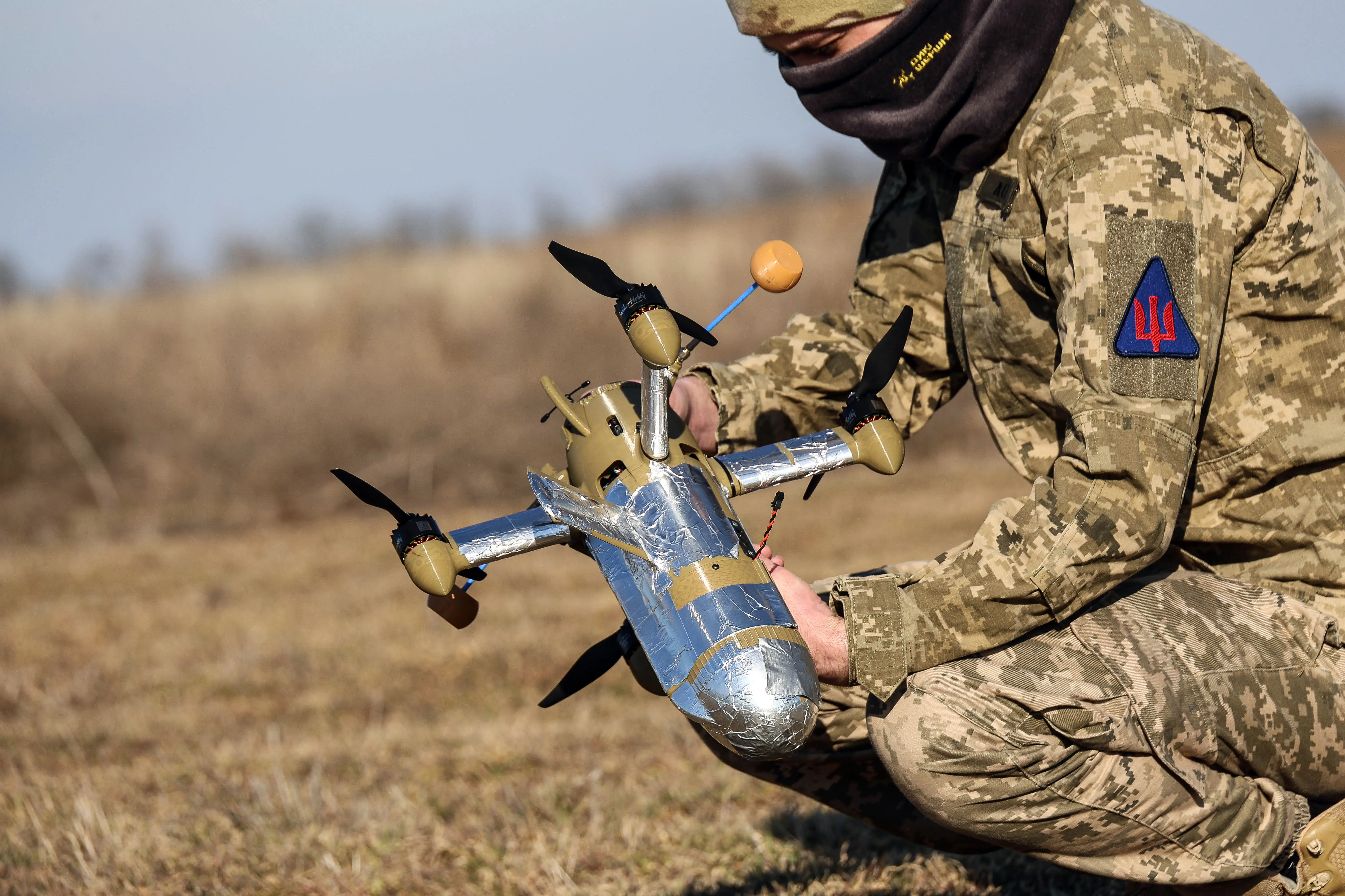 A man in camoufage squats in a field looking at a silver interceptor drone