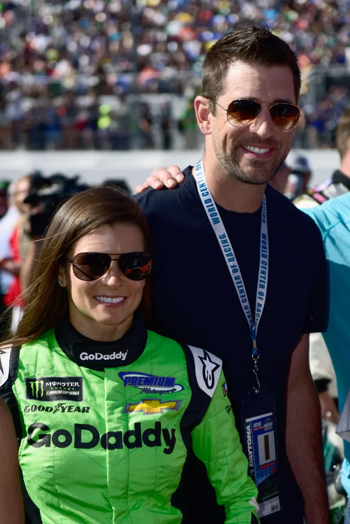 Danica Patrick in a green racing suit and sunglasses stands next to Aaron Rodgers in a dark blue shirt and sunglasses with his arm around her shoulder.