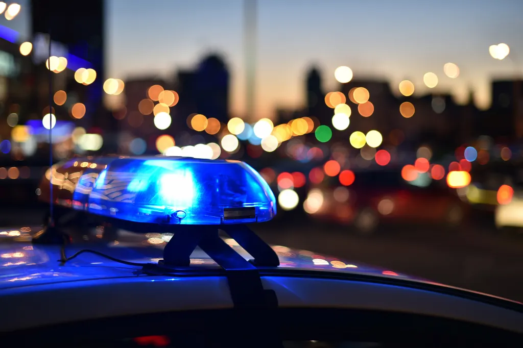 The blue lights of a police car flash at night with blurred city lights in the background.