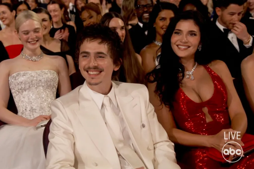 Timothée Chalamet in a white suit, Kylie Jenner, in a red gown, and Elle Fanning, behind them in a sparking silver gown, in the audience at the Academy Awards.