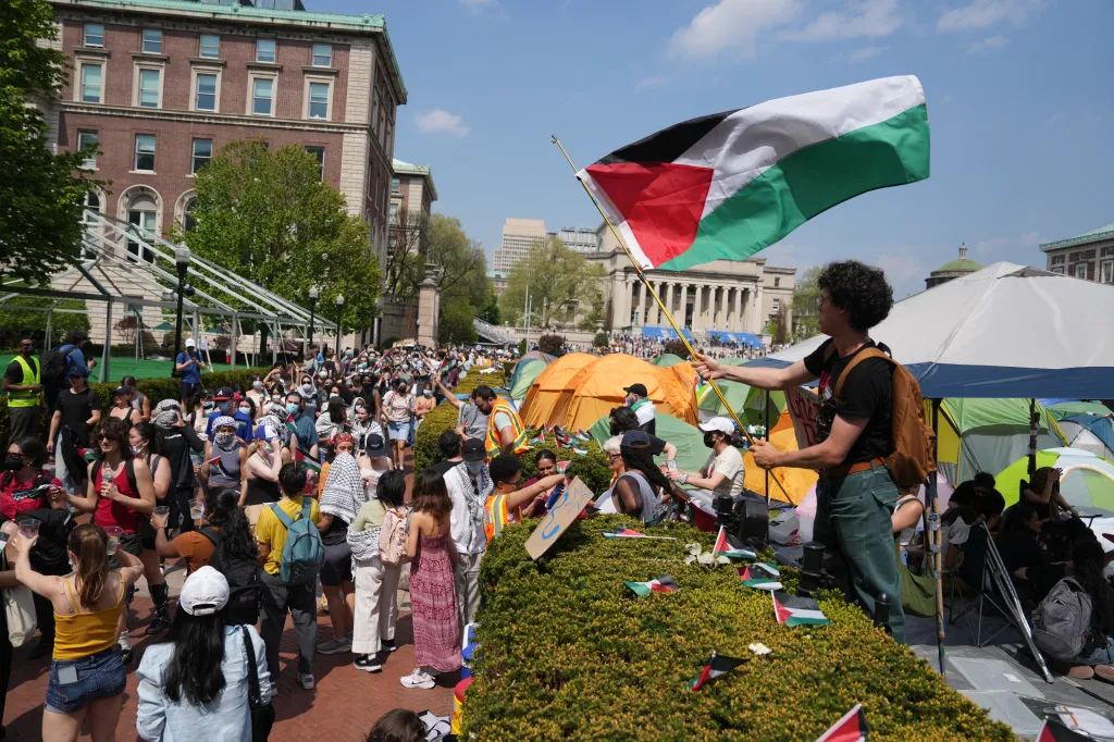 A man holds a Palestinian flag while standing among protesters, tents, and small Palestinian flags on a hedge in an encampment at Columbia University.