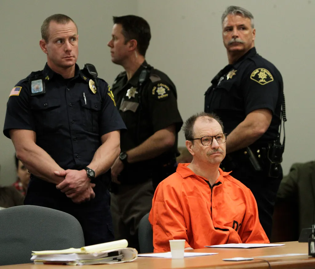 Gary Ridgway, the Green River Killer, in an orange jumpsuit at a table in a courtroom, flanked by three law enforcement officers.