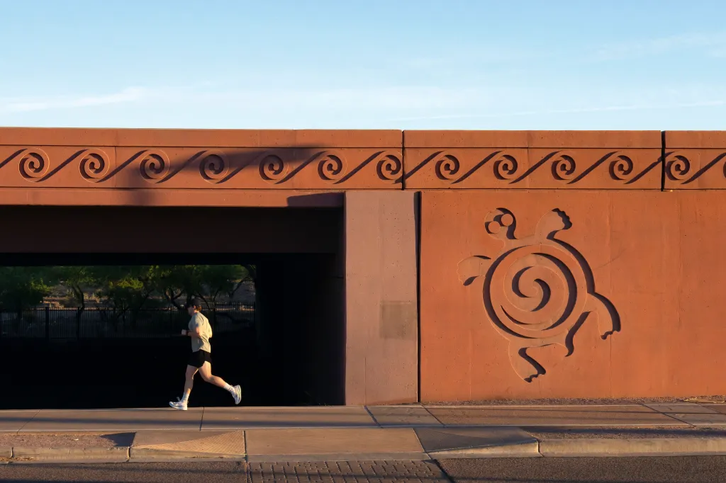 A person running on a sidewalk next to a reddish-brown underpass decorated with a turtle figure and a repeating wave pattern.
