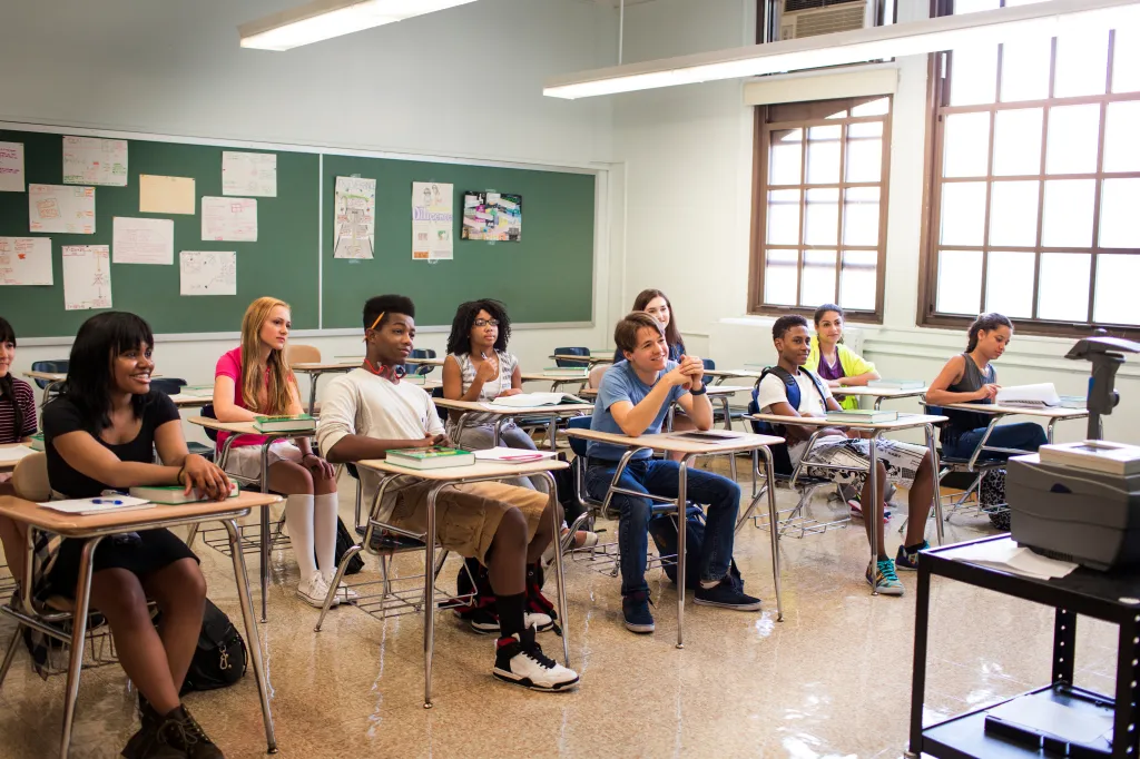 Students of diverse backgrounds sit at desks in a classroom with a green chalkboard.