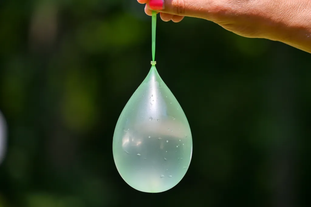 A hand with pink fingernails holds a translucent green water balloon with a green clip, against a dark green background.