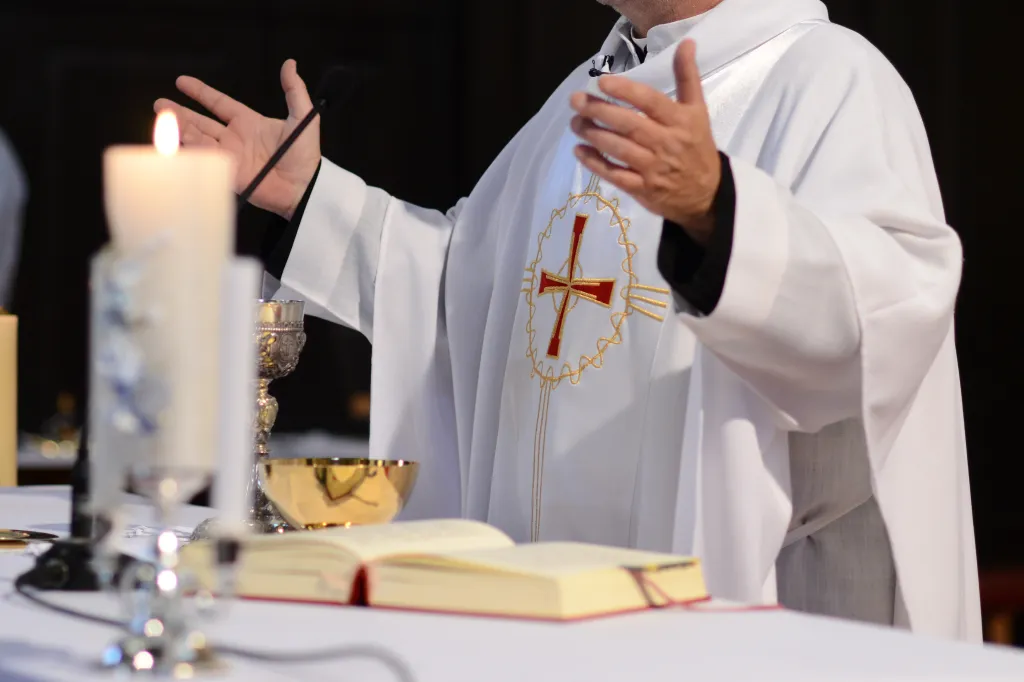 A priest wearing a white robe with a red and gold cross at the chest, stands at an altar with arms outstretched, next to a lit candle, chalice, and open book.
