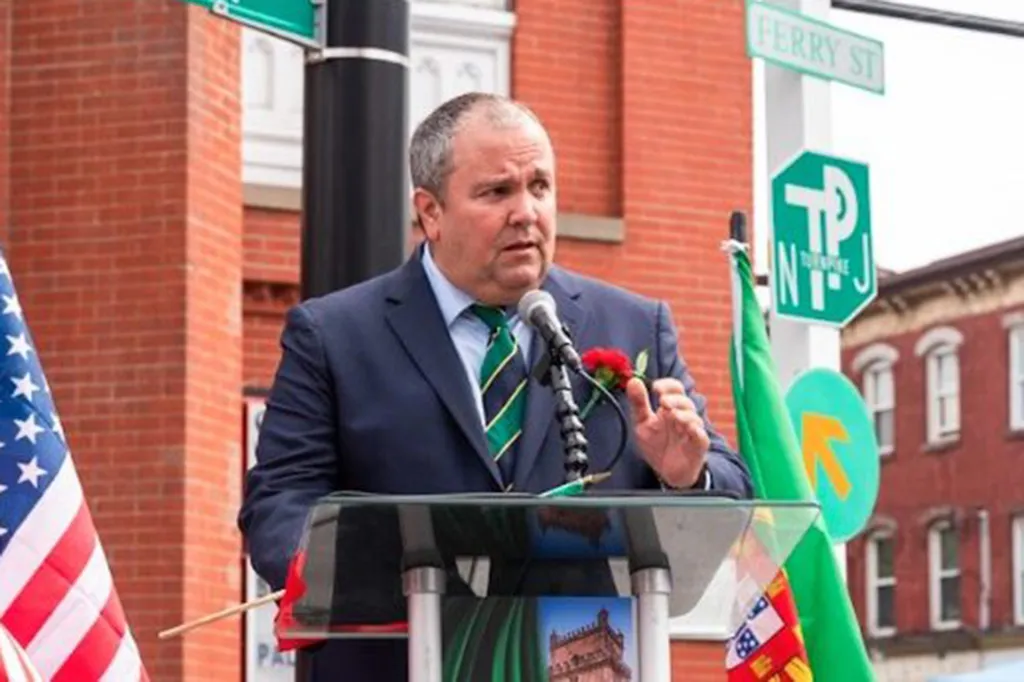 Albert Coutinho speaking at a podium with an American flag and a Portuguese flag in the foreground.