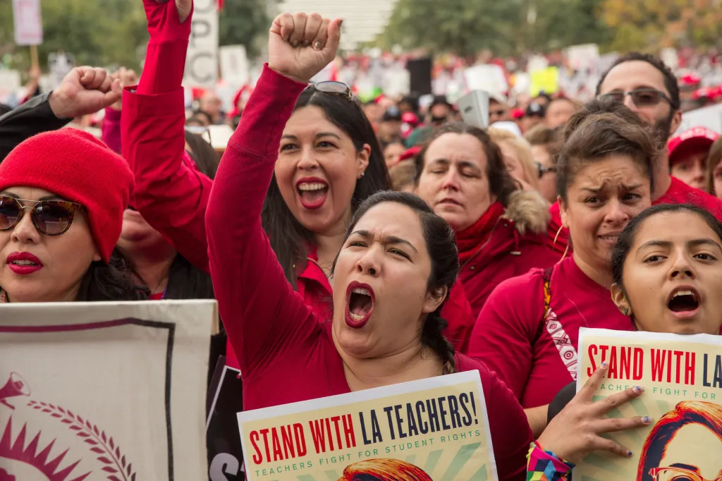 A crowd of teachers and supporters, many wearing red, protest with raised fists and signs that read 