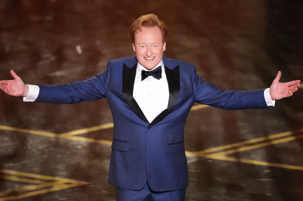 Conan O'Brien smiling with arms outstretched in a blue tuxedo.