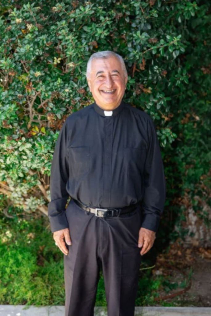 Emmanuel Shaleta, a smiling man in a black clerical shirt, standing in front of green foliage.