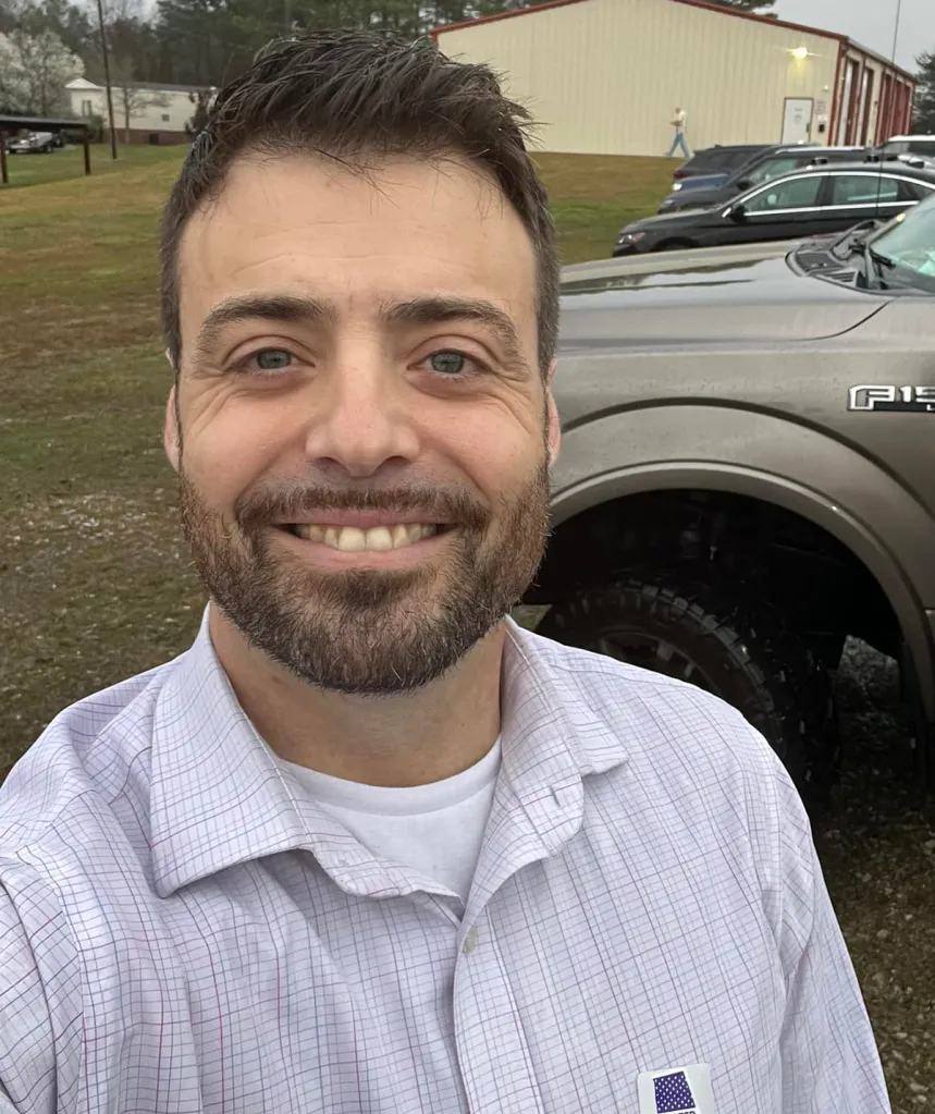 A man in a plaid shirt with a beard and a bright smile.