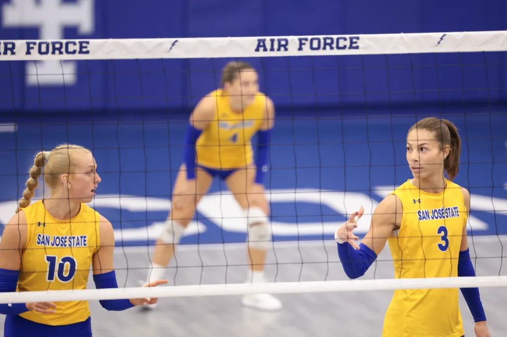 Three volleyball players in yellow and blue uniforms stand on a court behind a net.