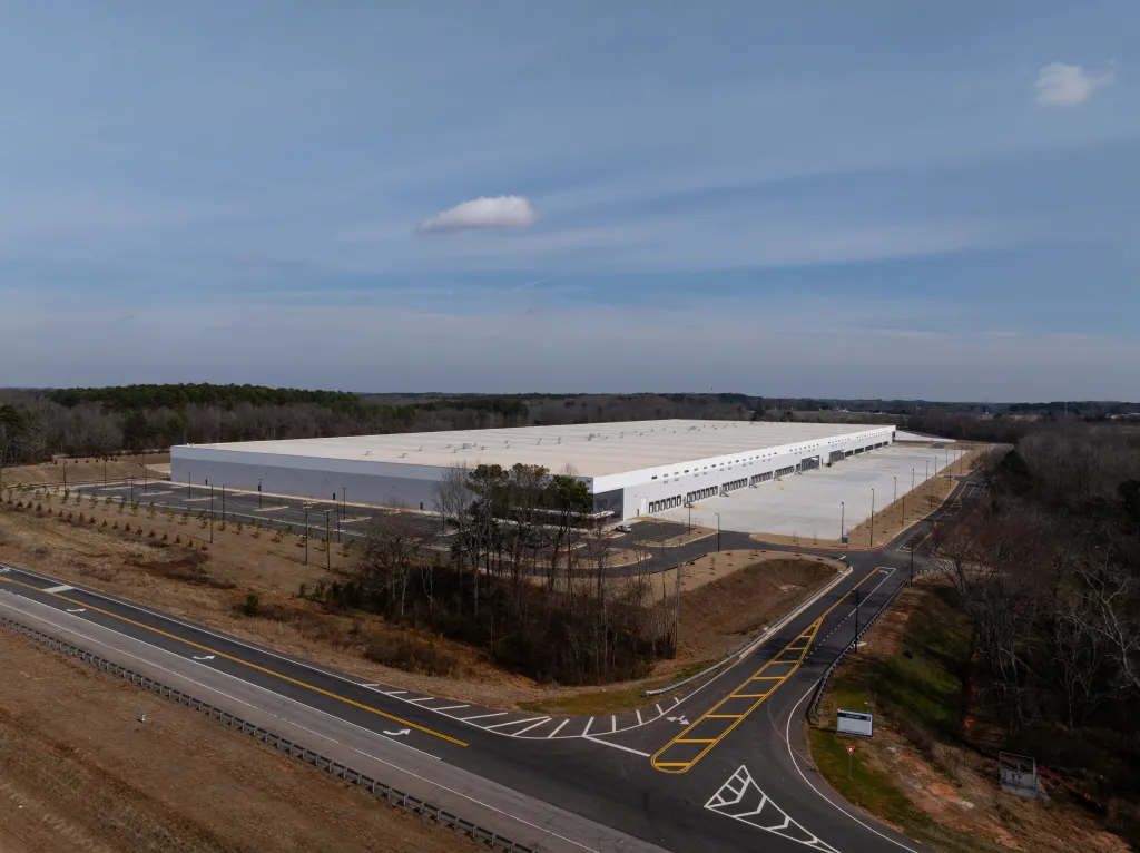 Aerial view of a large, long warehouse building with many loading docks, surrounded by bare trees, empty parking lots, and roads, under a cloudy sky.