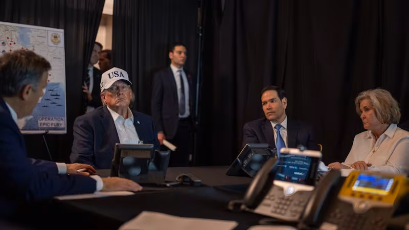 Trump in a white USA trucker hat in a darkened room with a map behind him and aides around him.