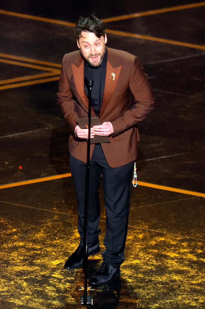 Man in a brown suit speaking into a microphone on stage.