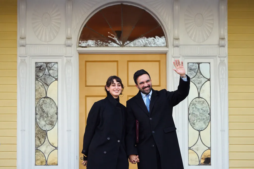 Mayor Zohran Mamdani waves to the camera while standing next to another person in front of a yellow building with white trim.