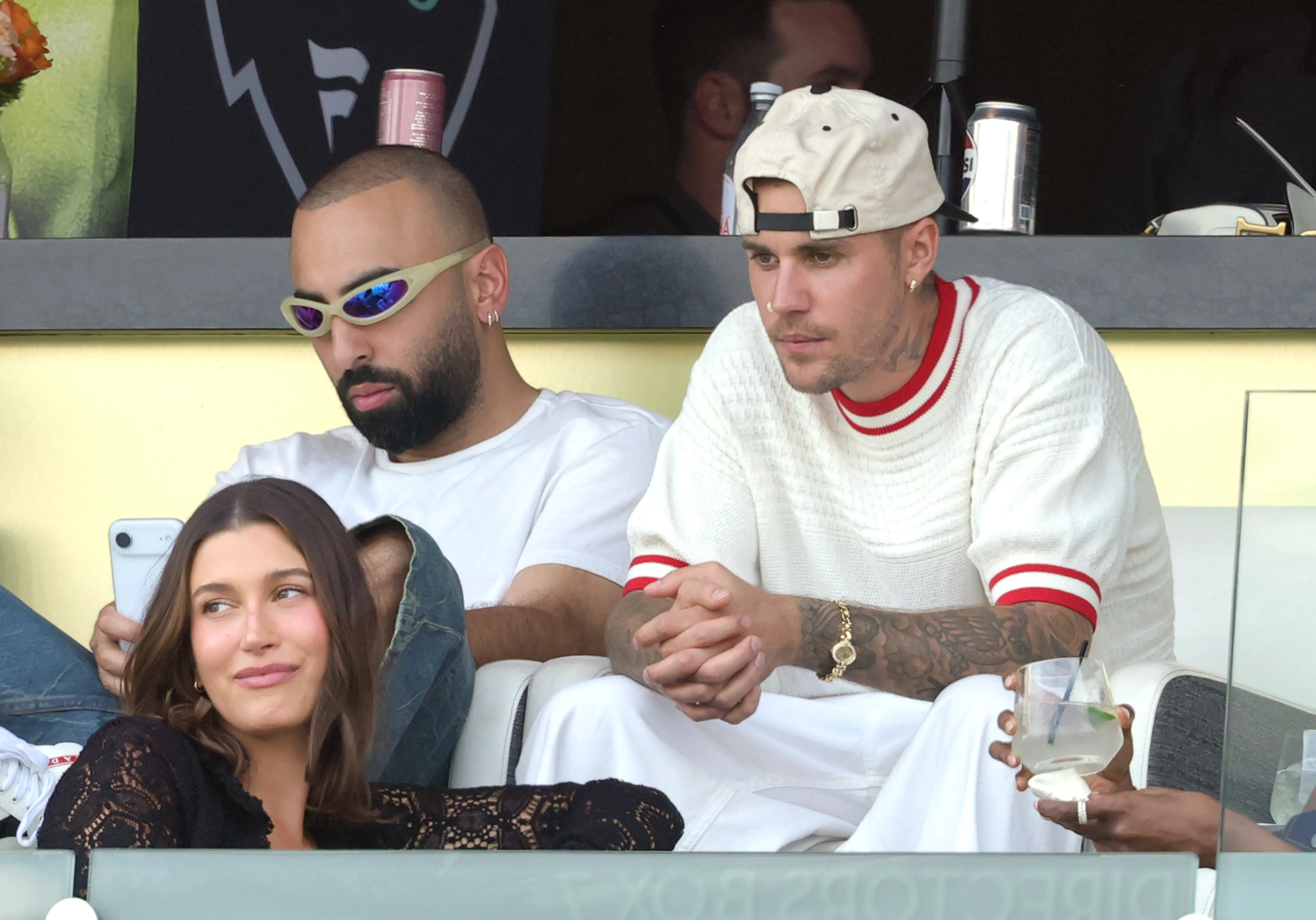 Hailey Bieber, wearing a black lace shirt, looks to the right, sitting in front of a man in sunglasses and a man in a white sweater and a backward baseball cap, at the Fanatics Flag Football Classic.