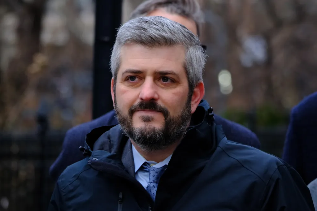 Man with gray hair and beard wearing a navy blue jacket and blue tie.