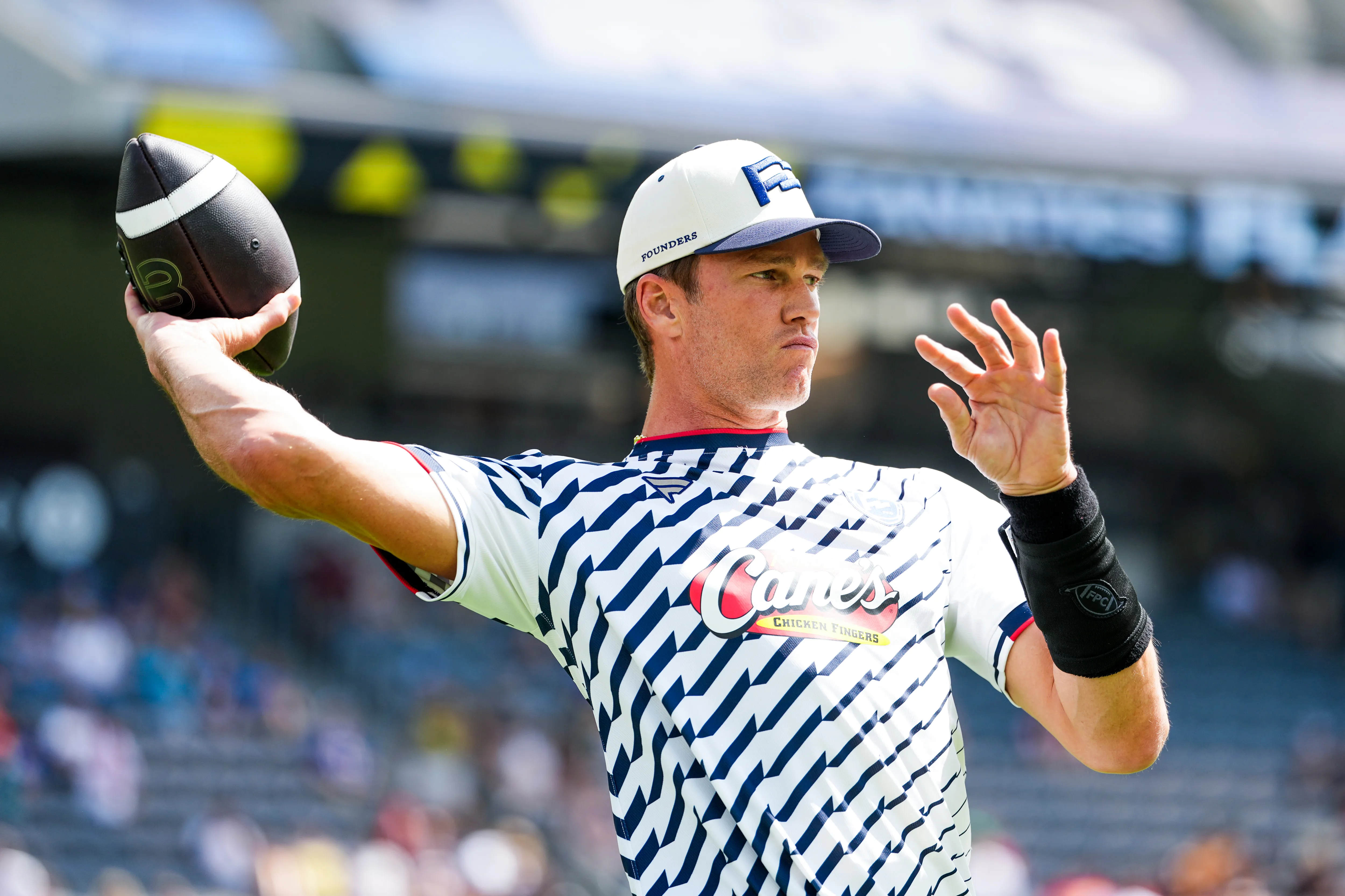 A man wearing a white and blue patterned shirt and a white baseball cap holds a football in his right hand, preparing to throw.