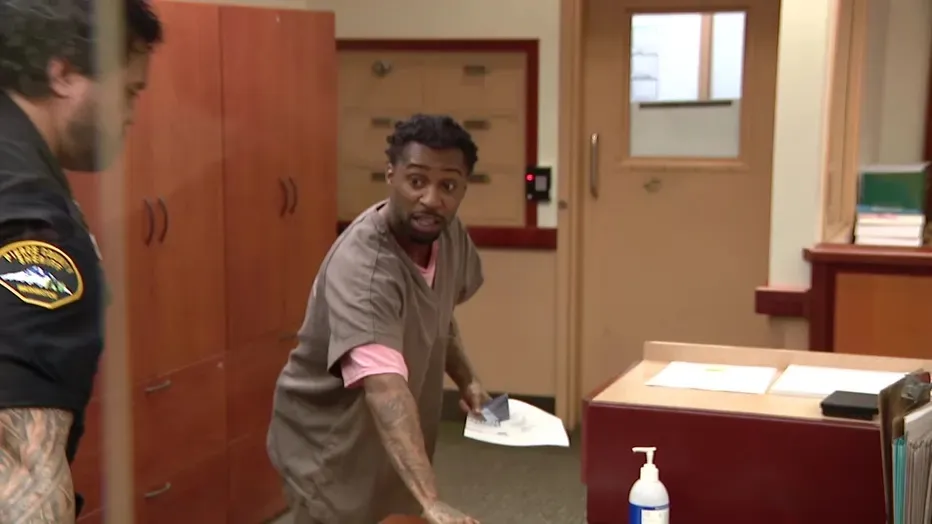 Majeed Guerry in a gray uniform with a pink shirt underneath, looking back as a man in a police uniform stands next to him.