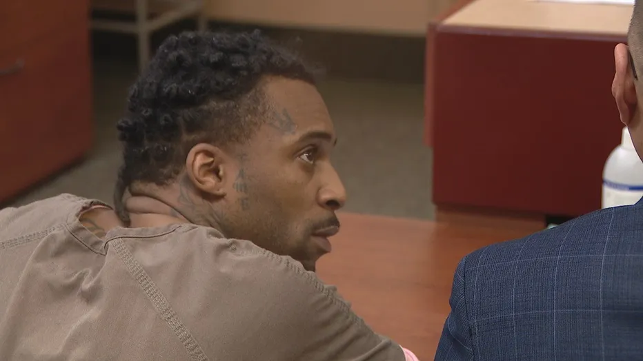 Majeed Guerry facing right, wearing a light brown shirt, with tattoos on his face and neck.