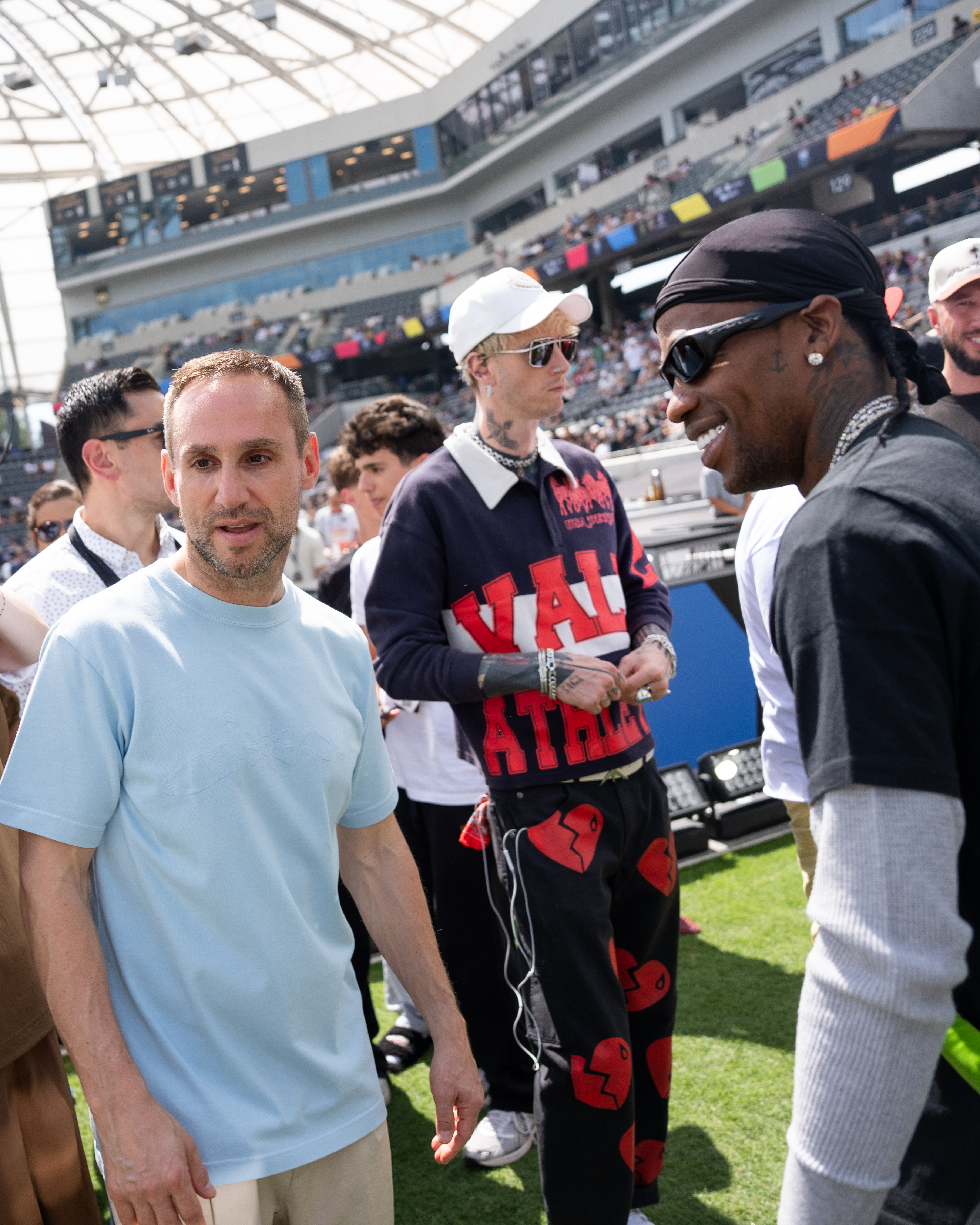 Machine Gun Kelly, a person in a durag and sunglasses, and another person on a sunny day in a stadium.