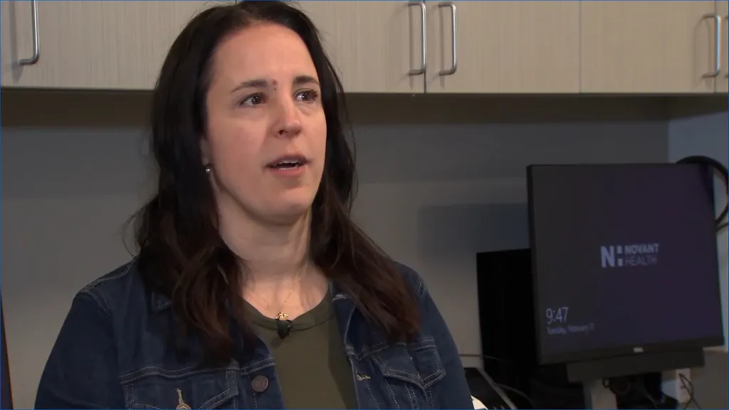A woman with long dark hair, wearing a denim jacket over a green shirt, speaks while a Novant Health computer monitor is visible in the background.