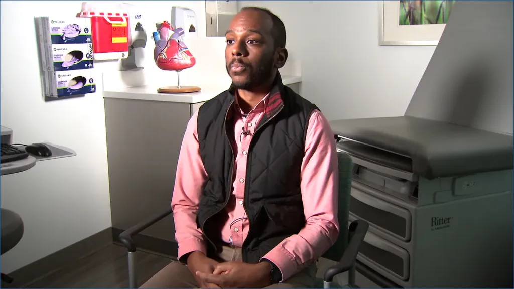 A man sits in a doctor's office, facing right, with a model of a human heart on a stand behind him.