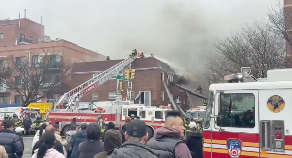 Firefighters battle a building fire with thick smoke billowing from the roof, seen from a crowd of onlookers.