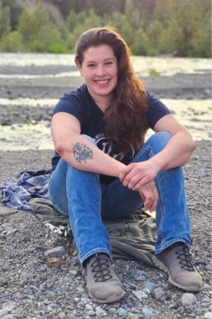 A woman with long brown hair sits on a blanket on a rocky riverbank, smiling at the camera.