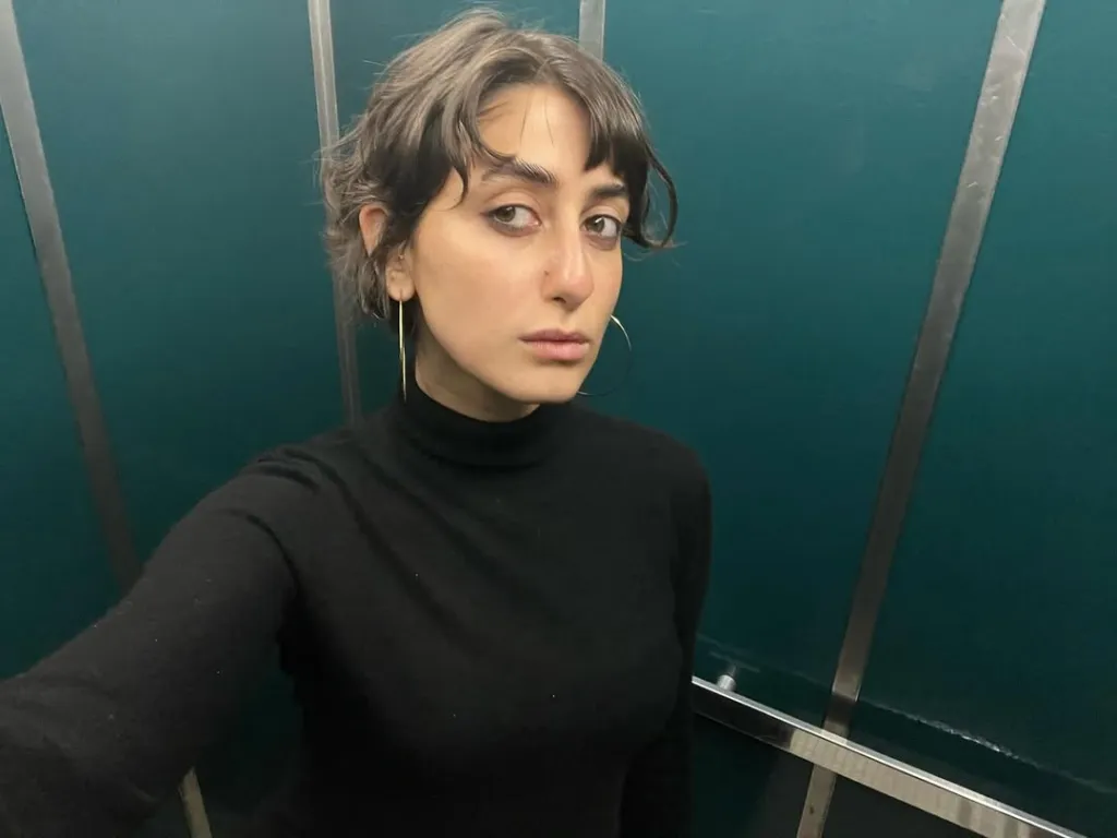 A young woman with short curly hair, bangs, and a black turtleneck, wearing long gold earrings and a hoop earring, stands in front of a teal wall with silver strips.