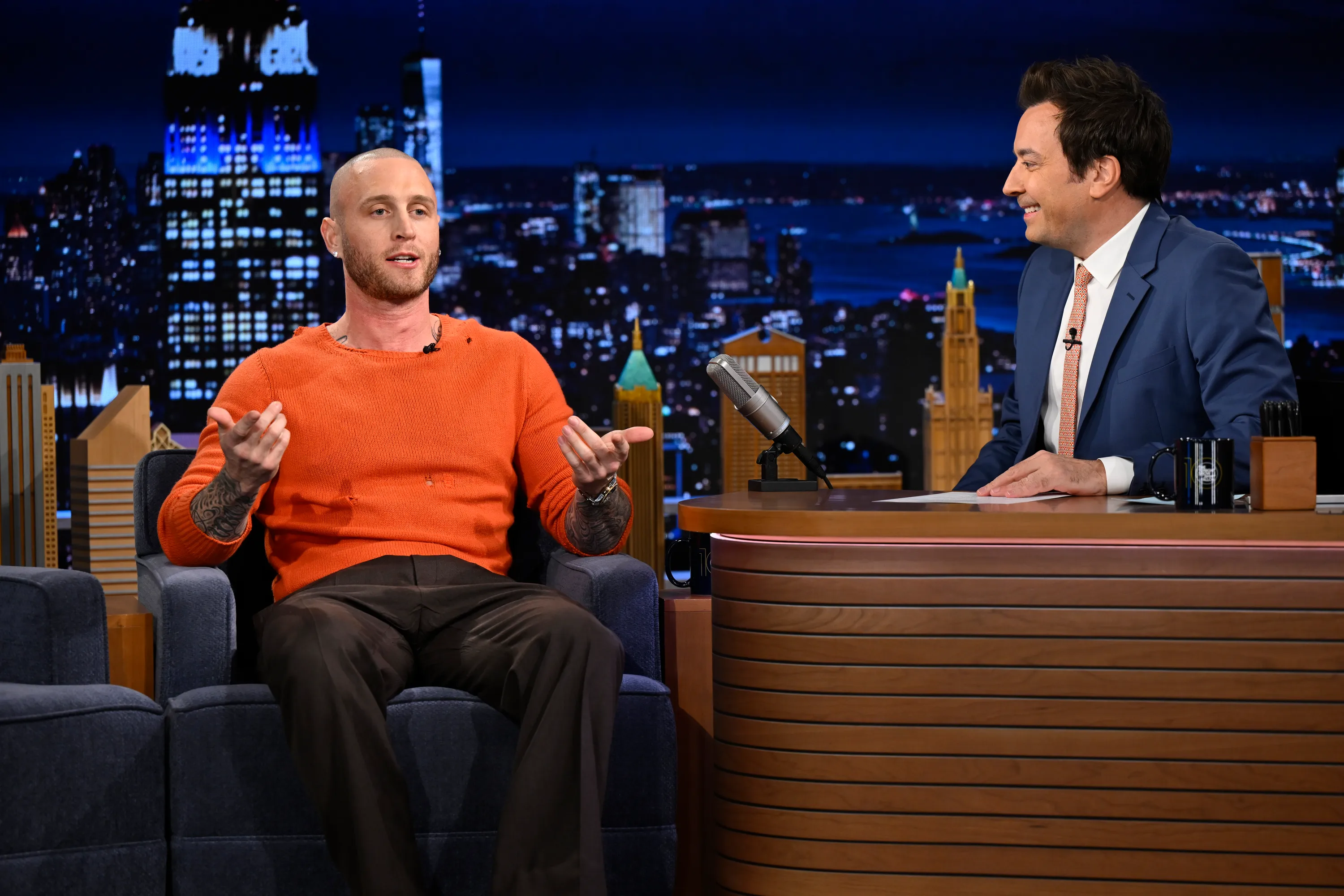Chet Hanks being interviewed by Jimmy Fallon on a talk show set with a city skyline backdrop.