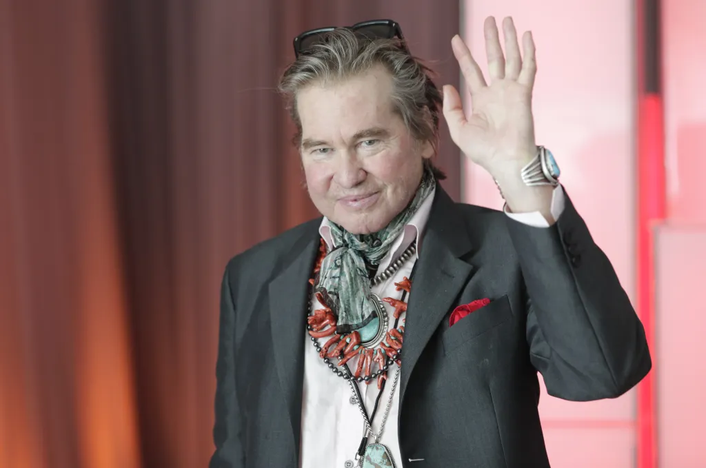 Val Kilmer waves while wearing a suit, scarf, and turquoise jewelry.