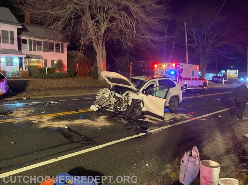 A white car with a severely damaged front end, an open hood, and an open driver's side door on a road at night, with an ambulance in the background.