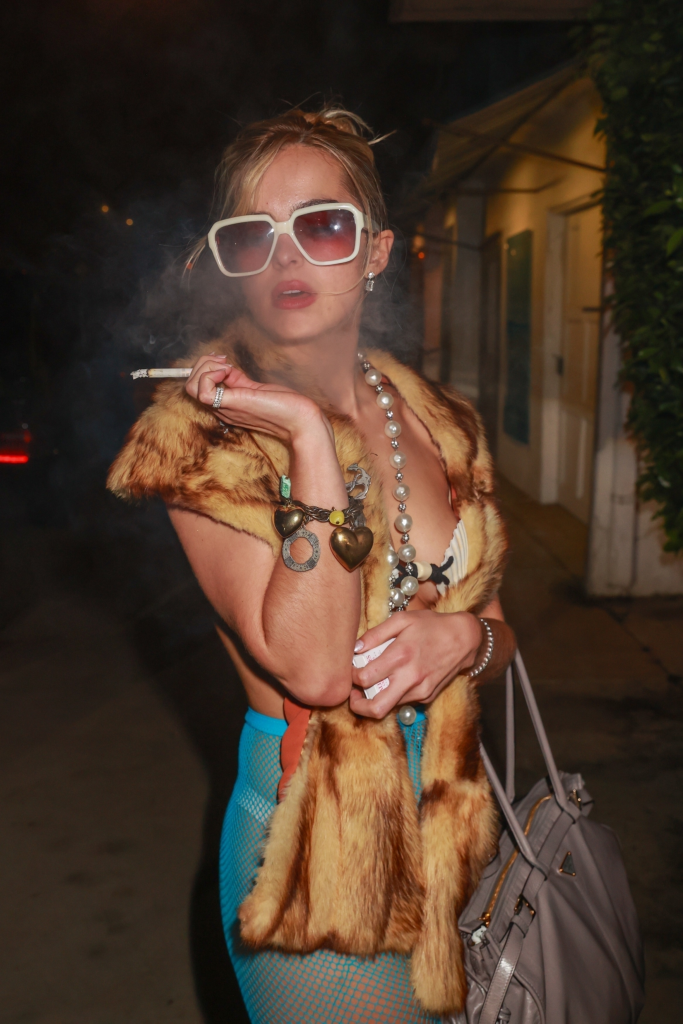 Addison Rae smoking a cigarette, wearing sunglasses, a fur stole, a pearl necklace, a bracelet with heart-shaped charms, and a blue fishnet dress.