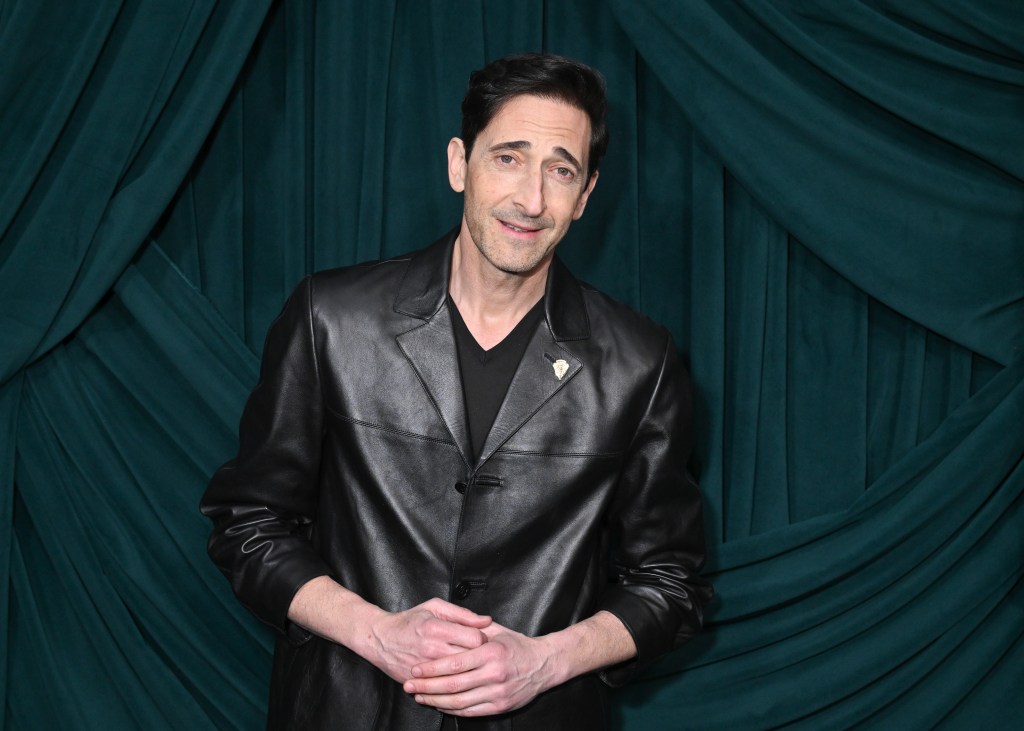 Adrien Brody in a black leather jacket, hands clasped, against a dark teal pleated backdrop.
