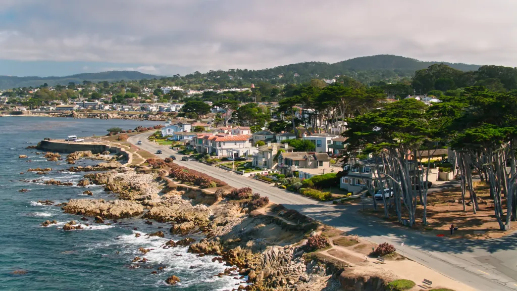 Aerial view of the Pacific Grove coastline, showing houses, a coastal road, and Cypress trees overlooking the ocean.