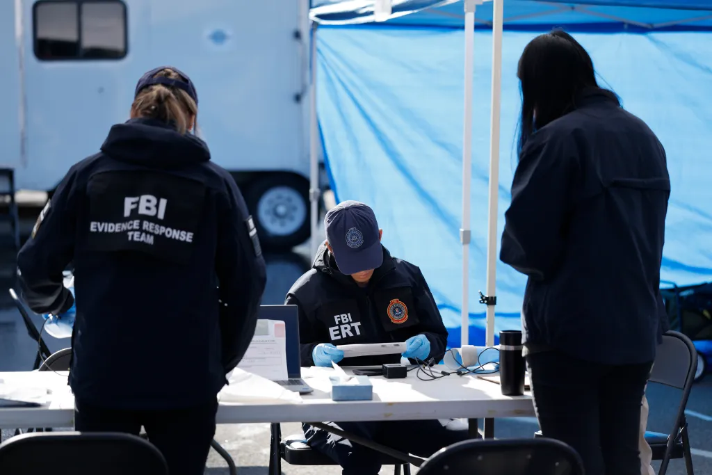 FBI agents work at a field command center, with one agent wearing gloves and reviewing documents at a table.