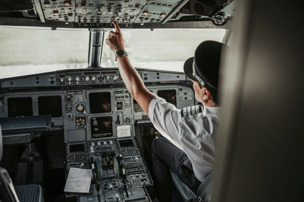 A mature pilot in uniform and cap inside a cockpit adjusts a switch on the overhead control panel.