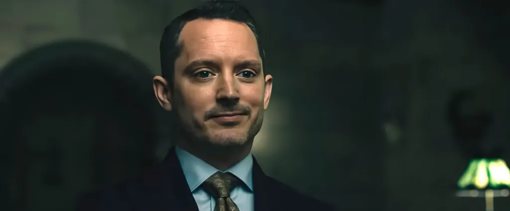Elijah Wood in a dark suit, light blue shirt, and patterned tie with a slight smile.