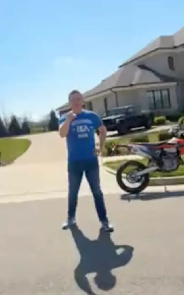 Alan Ritchson stands outside a large house, a black pickup truck and a dirt bike visible behind him.