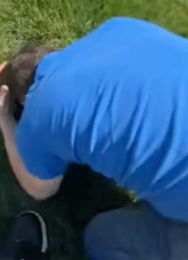 Alan Ritchson hunches over on a patch of grass, wearing a blue shirt.