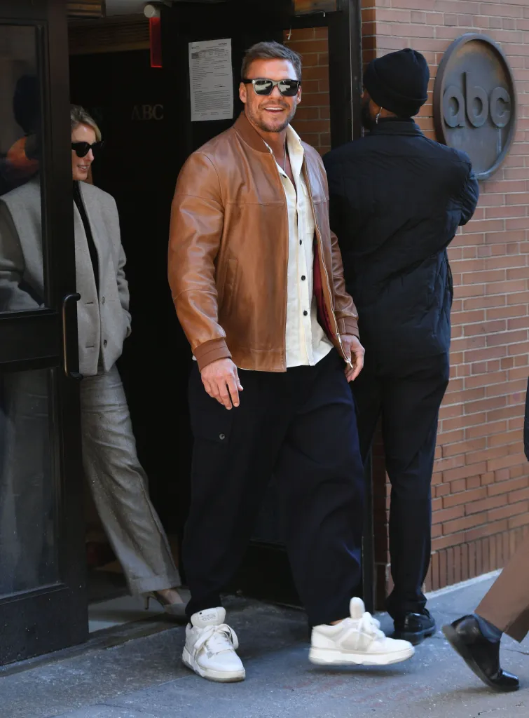 Alan Ritchson wearing a brown leather jacket and sunglasses.