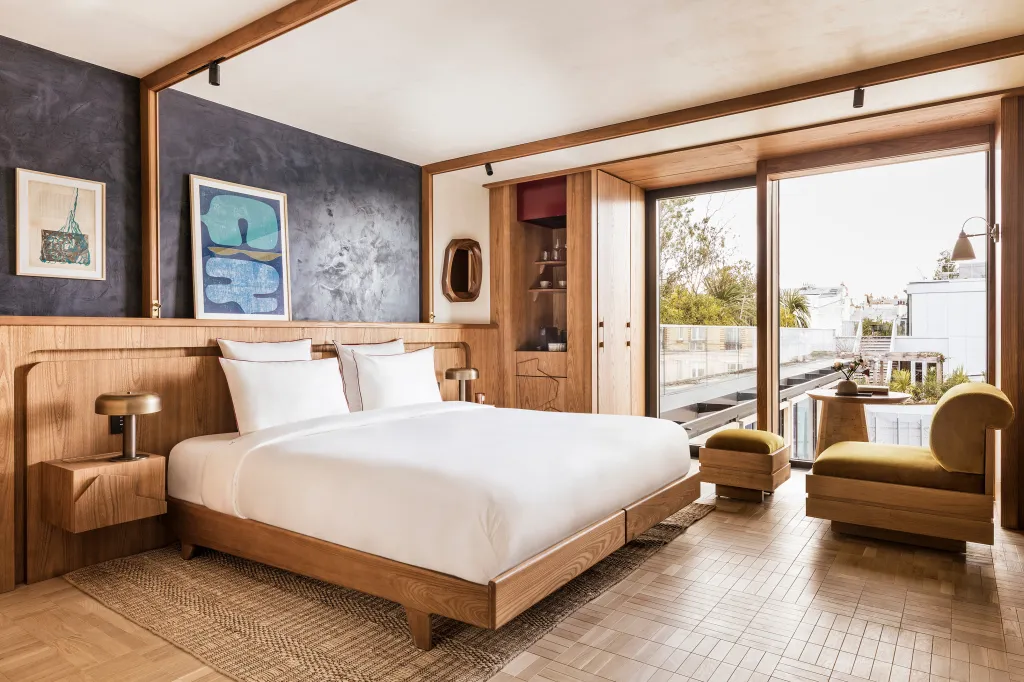 A modern bedroom with a large bed against a dark accent wall, two abstract art prints, a built-in wooden cabinet, and a sitting area with a large window.