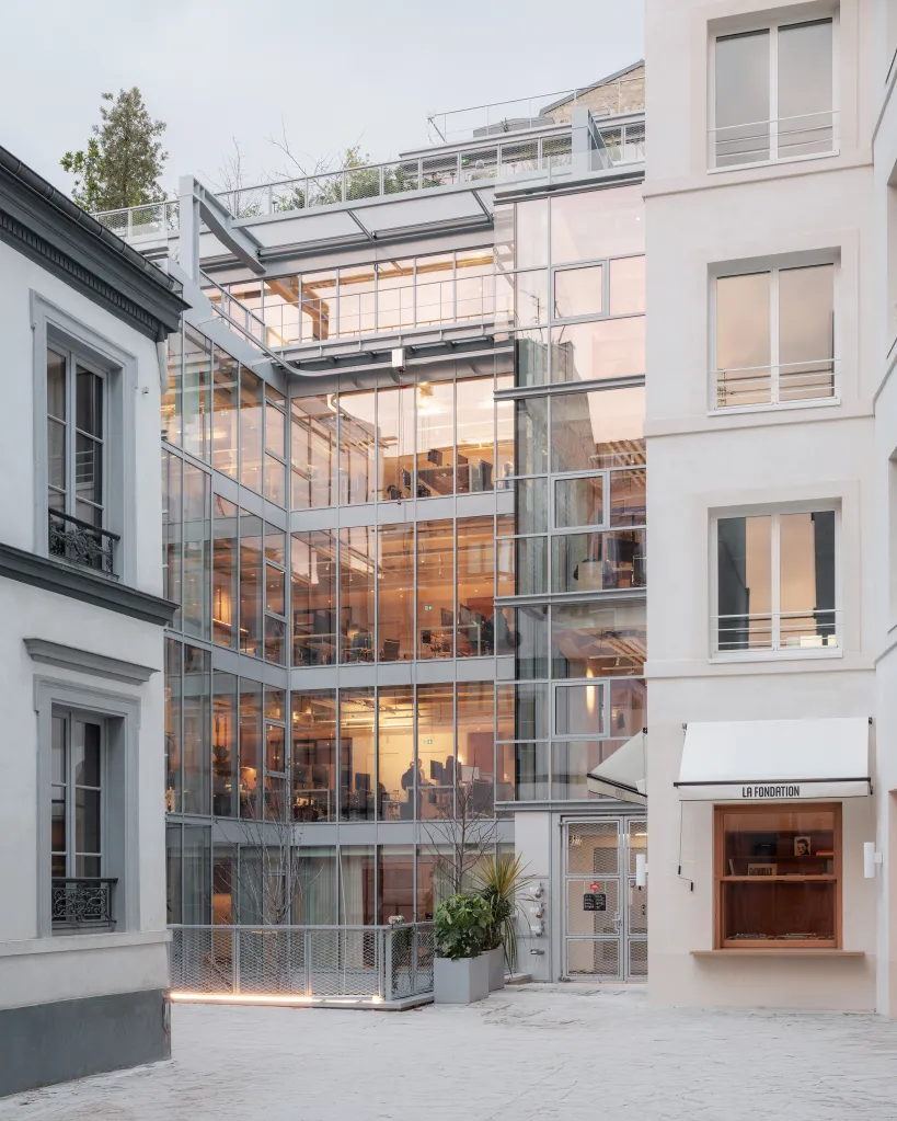 La Fondation, a modern building with a glass facade reflecting sunset hues, surrounded by older, light-colored buildings with traditional windows, all within a Parisian courtyard.