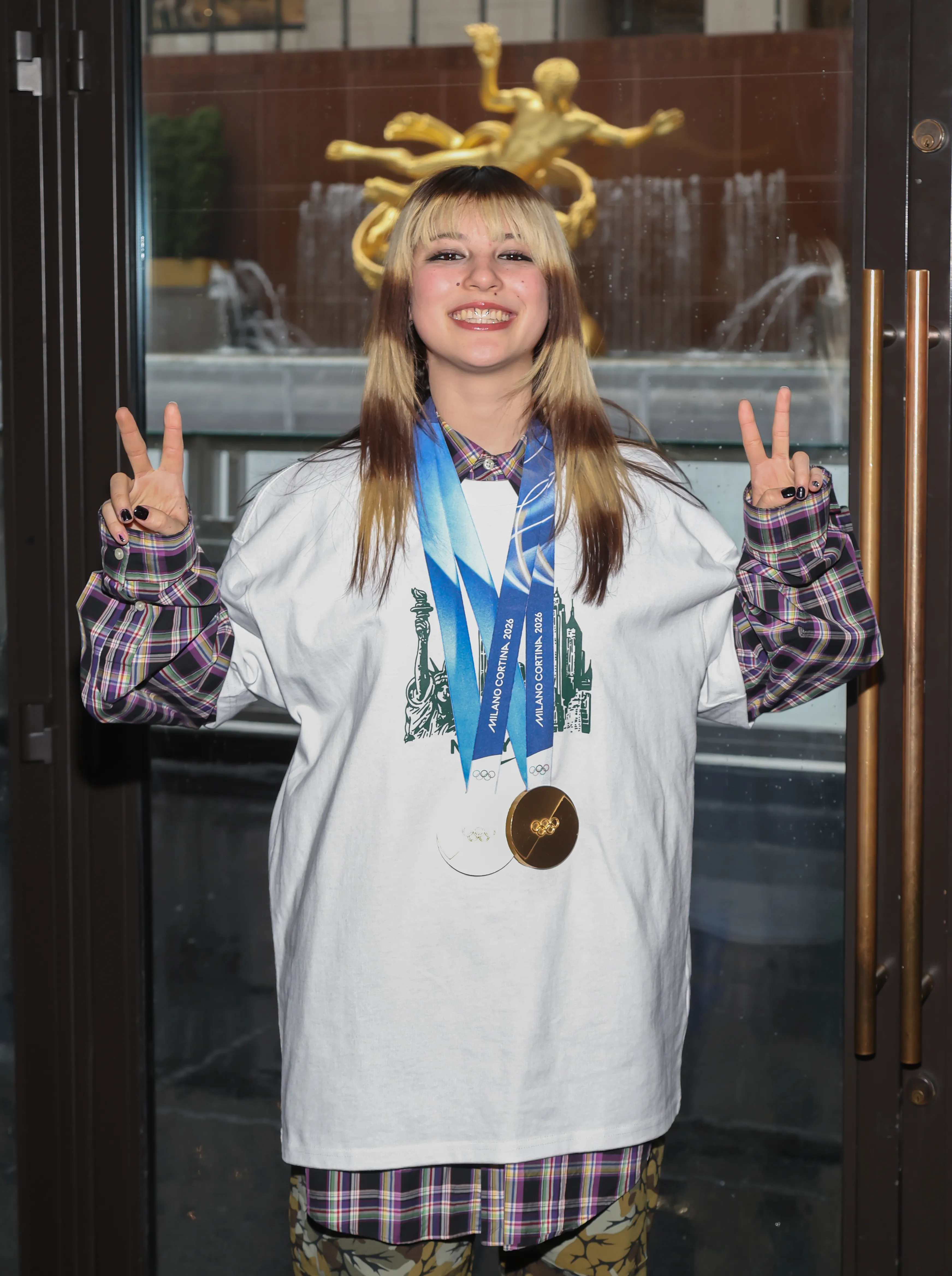 Alysa Liu wearing two Olympic medals, making a peace sign with both hands at Rockefeller Center.