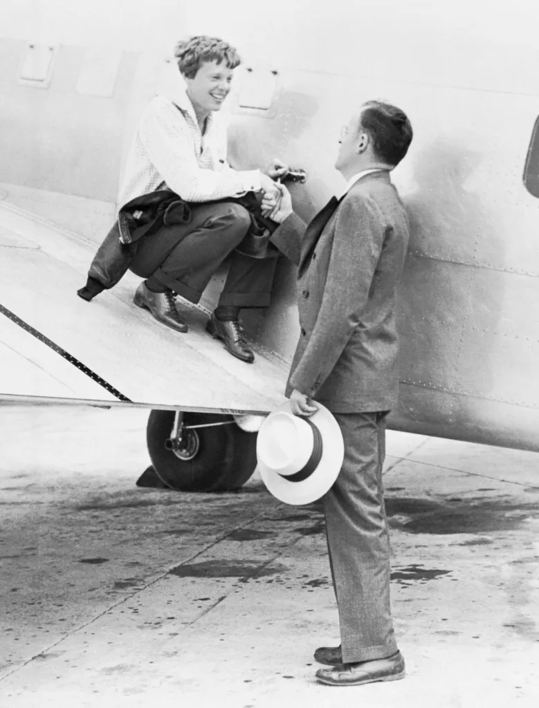 Amelia Earhart shaking hands with her husband George Palmer Putnam while sitting on the wing of a plane.